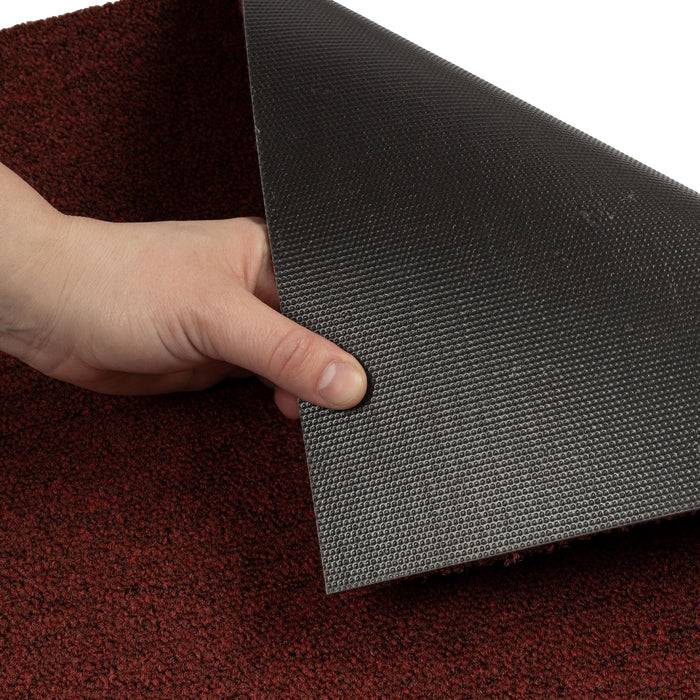 Eine Hand hebt die Ecke einer roten, strukturierten Matte an, um ihre schwarze, rutschfeste Rückseite zu zeigen, und demonstriert damit die Flexibilität und Griffigkeit der Matte.