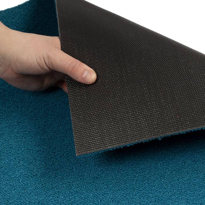 Eine Hand hebt die Kante eines blaugrünen Teppichs an und offenbart seine rutschfeste schwarze Rückseite, die das Sicherheitsmerkmal des Teppichs hervorhebt.