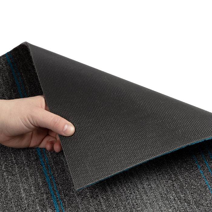 Eine Hand hebt die Ecke eines grau und schwarz strukturierten Teppichs an und gibt damit eine rutschfeste Unterlage frei. Blaue Streifen verlaufen entlang der Teppichoberfläche.