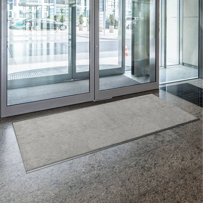 Ein großer Glaseingang mit Spiegelungen führt zu einer modernen Gebäudehalle. Eine graue Fußmatte liegt auf poliertem, dunkelm Marmorboden.