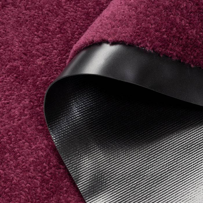 „Nahaufnahme eines dunkelroten strukturierten Teppichs mit einer gekrümmten Kante, die eine glatte schwarze Unterseite zeigt.