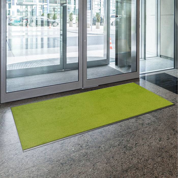 Eine hellgrüne Matte auf einem polierten Boden neben großen Glastüren, die nach außen zum Eingangsbereich eines Gebäudes führen.