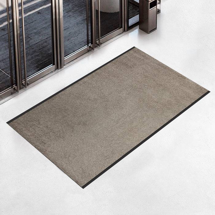 Eine graue Bodenmatte mit schwarzem Rand liegt auf einem hellgrauen, polierten Boden in der Nähe großer Glastüren und einer Wand mit Metallleisten.