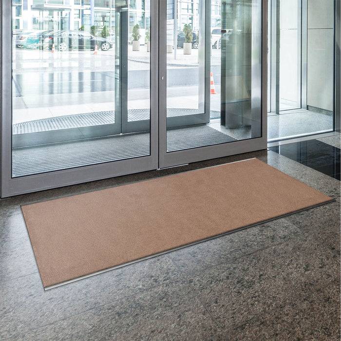 Eine braune Fußmatte liegt auf dem grauen Steinboden vor großen Glastüren in der Eingangshalle eines Bürogebäudes und bietet einen willkommenen Eingang.