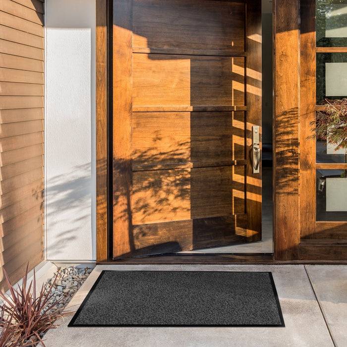 Eine moderne Holz-Eingangstür mit einem großen Griff, umgeben von einer hellfarbenen Fassade. Sonnenlicht wirft Schatten auf die Tür. Ein dunkles Türvorleger liegt davor.