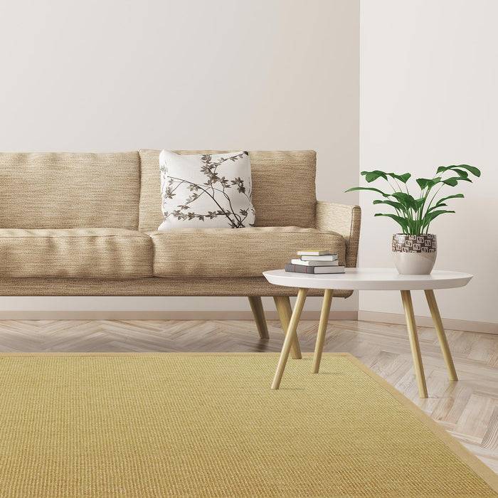 Ein beiges Sofa mit einem gemusterten Kissen lehnt an einer hellen Wand. Ein weißer runder Tisch mit einer Topfpflanze steht in der Nähe des Sofas auf einem Holzboden.