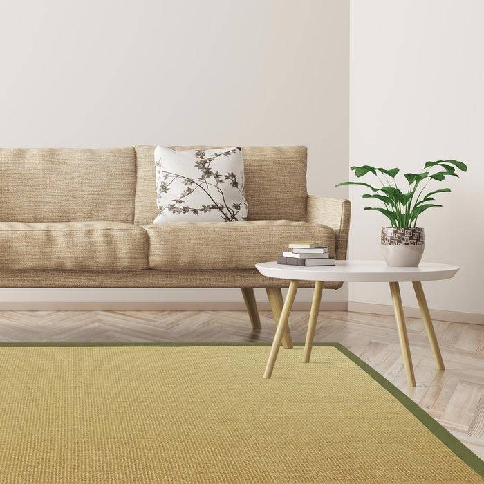 Ein beigefarbenes Sofa mit einem gemusterten Kissen lehnt an einer weißen Wand. Ein kleiner Tisch mit einer Topfpflanze steht auf einem grün eingefassten Teppich in einem hell erleuchteten Raum.
