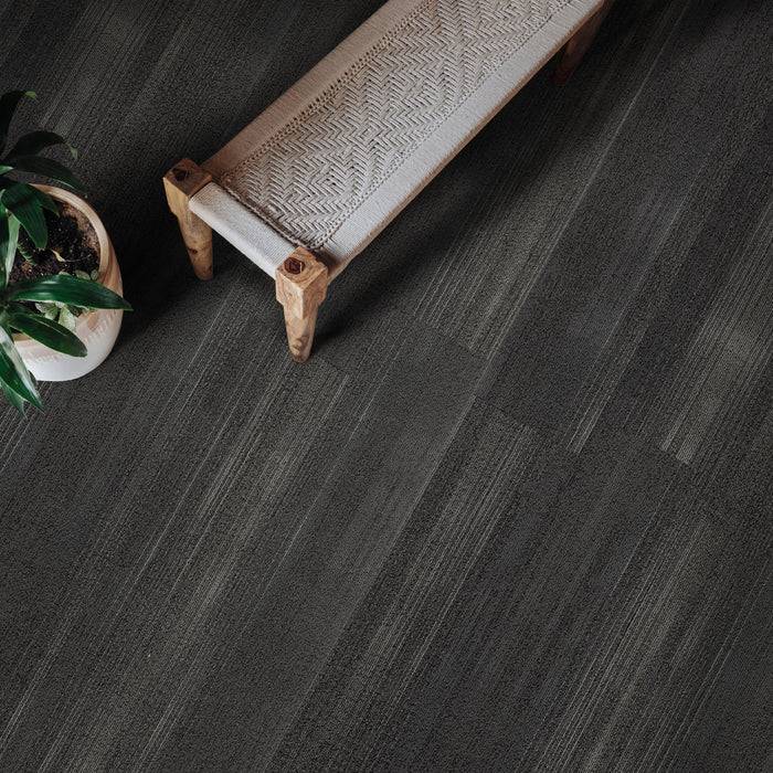 Eine minimalistische Szene mit einer gewebten Bank und einer getopften Pflanze auf einem strukturierten dunkelgrauen Bodenbelag, die eine moderne, ruhige Atmosphäre schafft.