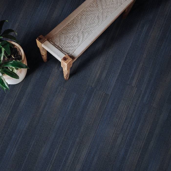 Eine Holzbank mit gewebtem Sitz steht auf einem dunklen Holzboden neben einer kleinen, üppig grünen Pflanze in einem Topf.