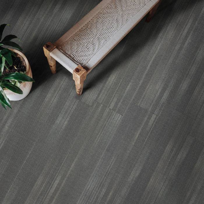 Eine lange Holzbank mit einem gemusterten Kissen steht auf einem grauem Fliesenboden. Eine Topfpflanze mit grünen Blättern steht neben der Bank.