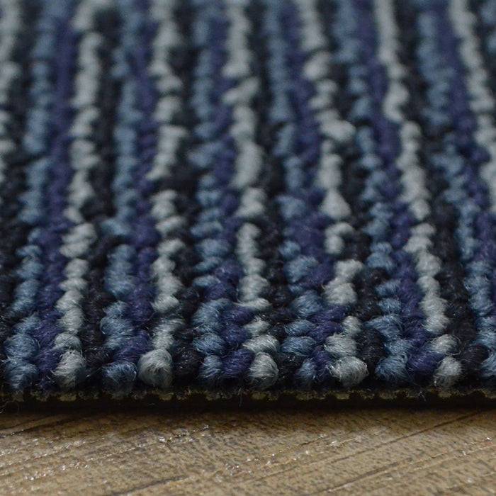 Nahaufnahme eines strukturierten, gestreiften Teppichs mit blauen, grauen und schwarzen Fäden, der auf einem Holzboden platziert ist.