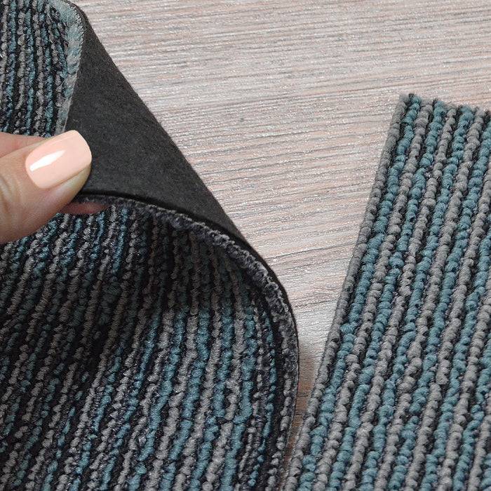 Eine Person hebt eine Ecke eines gestreiften, strukturierten Teppichs an, wodurch seine schwarze Rückseite auf einem hellen Holzboden sichtbar wird.