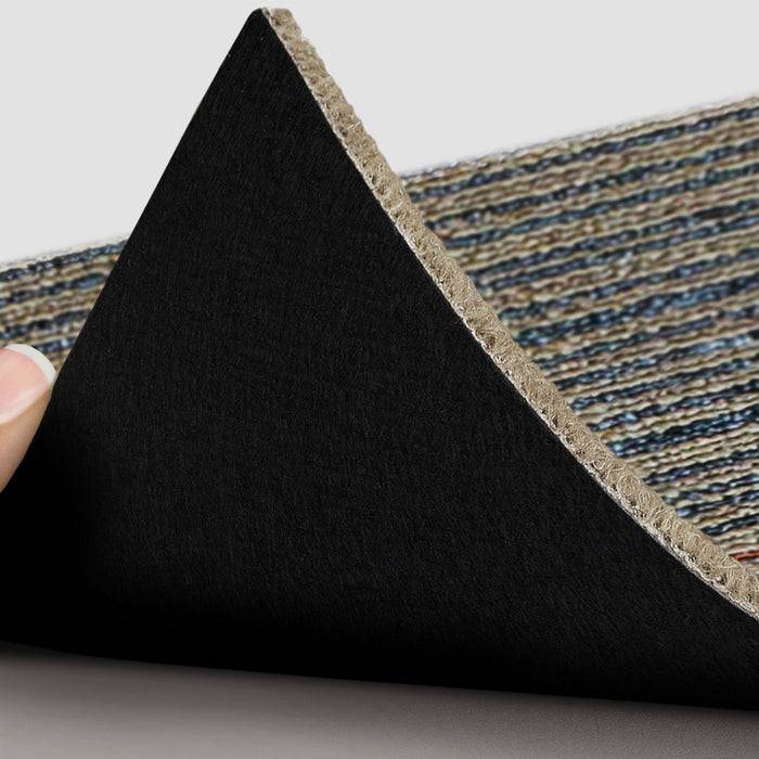 Eine Hand hebt die Ecke eines gestreiften, strukturierten Teppichs an und enthüllt dabei seine schwarze, rutschfeste Unterseite, was auf Haltbarkeit und Sicherheitsmerkmale hindeutet.