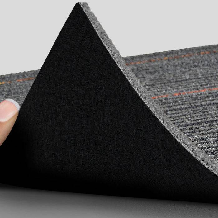 Eine Hand hebt die Kante eines schwarzen Teppichs mit sichtbarem Rücken an und zeigt eine Griffstruktur, die für rutschfeste Eigenschaften entwickelt wurde.