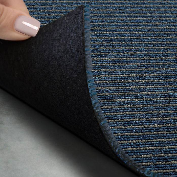 Eine Hand hebt eine Ecke eines dunkelblau gestreiften Teppichs an und gibt so dessen weiche, flexible Rückseite frei.