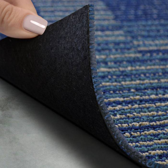 Eine Hand hebt die Ecke eines blauen Teppichs an und enthüllt dessen schwarze rutschfeste Rückseite auf einer grauen Oberfläche.