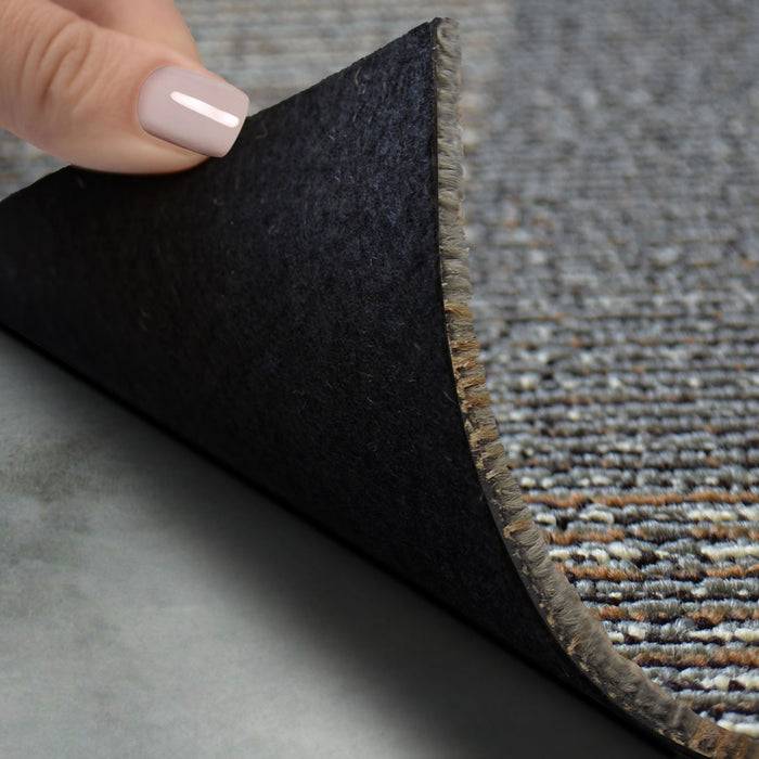 Eine Hand hebt eine Ecke eines Teppichs an und enthüllt eine schwarze rutschfeste Rückseite, die ihre Textur und Details auf einem grauen Boden zeigt.