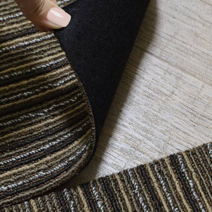 Eine Person hebt die Ecke eines schwarzrückigen, gestreiften Teppichs auf einem Holzboden an und enthüllt strukturierte Schichten.