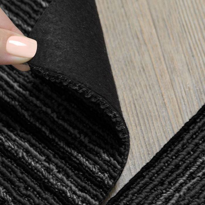 Eine Hand hebt die Ecke eines schwarzen gewebten Teppichs an und gibt eine strukturierte Oberfläche auf einem Holzboden frei.