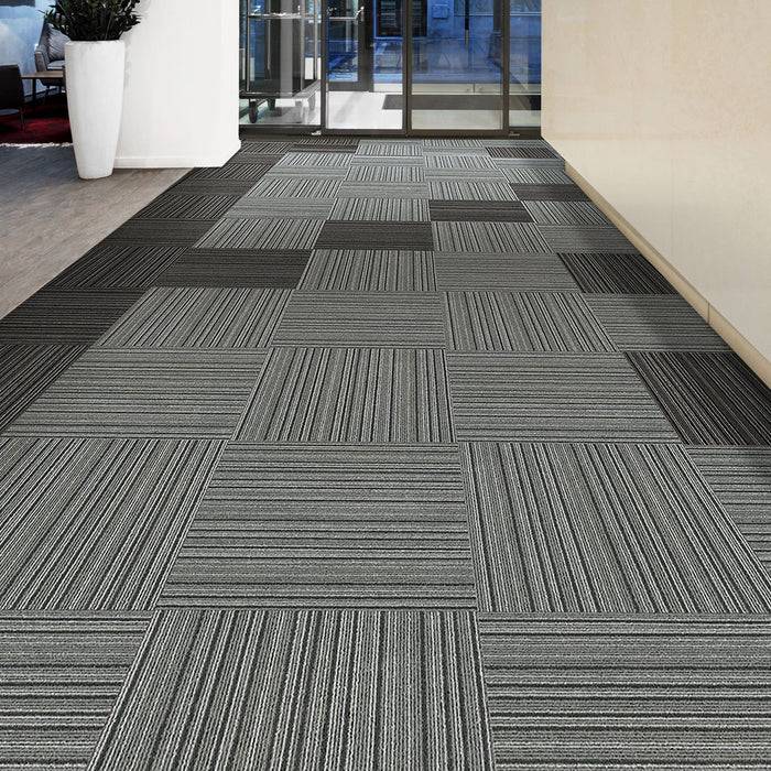 Ein Korridor mit einem modernen Teppich, der ein geometrisches Muster aus schwarzen, weißen und grauen Quadraten aufweist. Große Blumenkübel sind auf der linken Seite platziert.