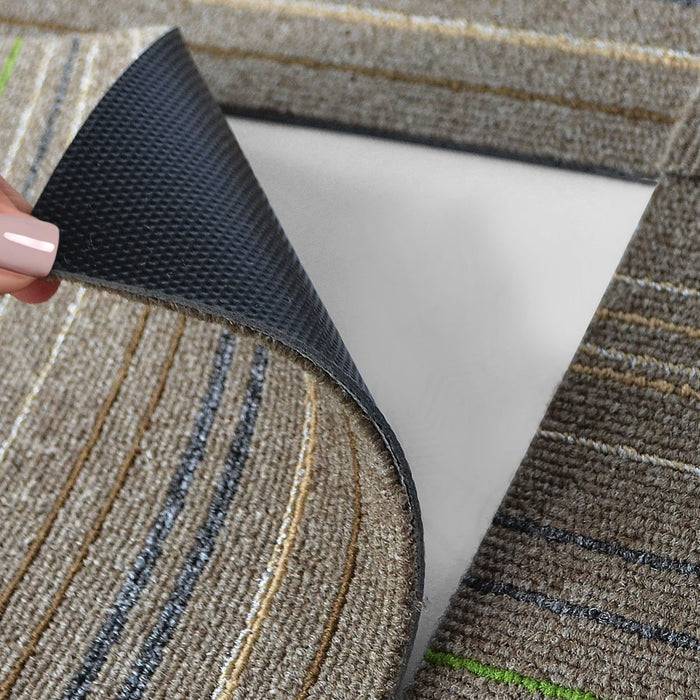 Eine Hand hebt eine Ecke eines gestreiften, strukturierten Teppichs an, um den darunter liegenden glatten, weißen Boden freizulegen und die Rückseite des Teppichs zu zeigen.