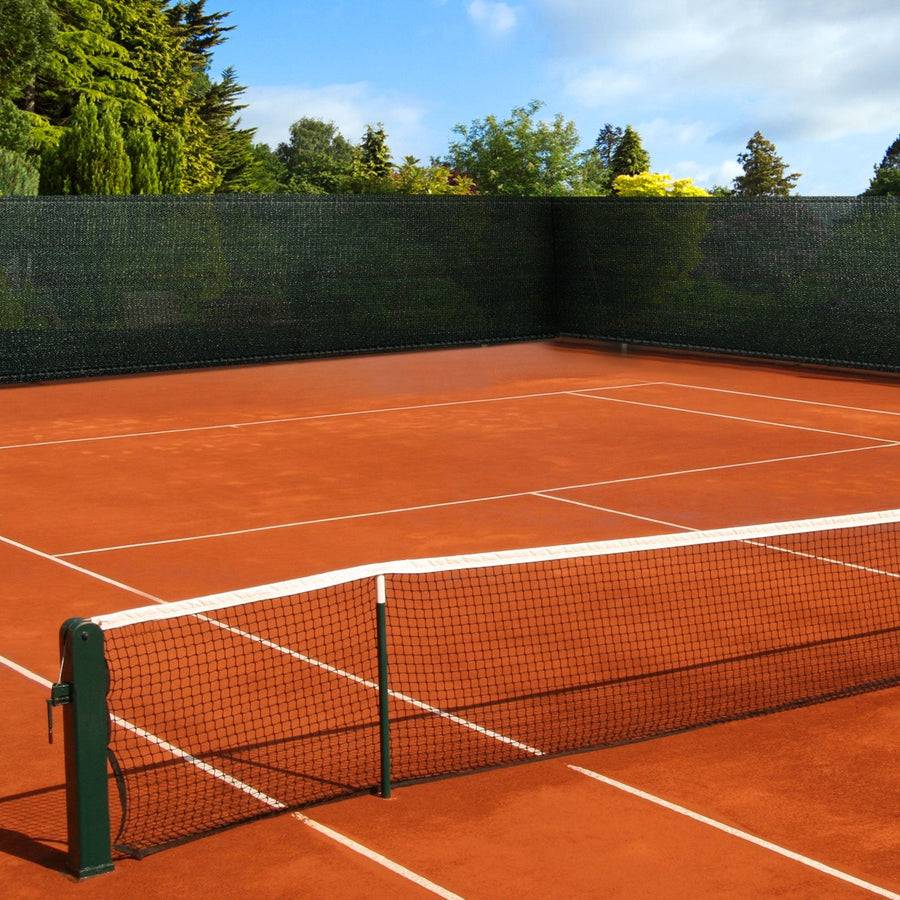 Ein Tennisplatz aus Lehm in einer Außenanlage, umgeben von grünen Bäumen und Büschen unter einem teilweise bewölkten Himmel.