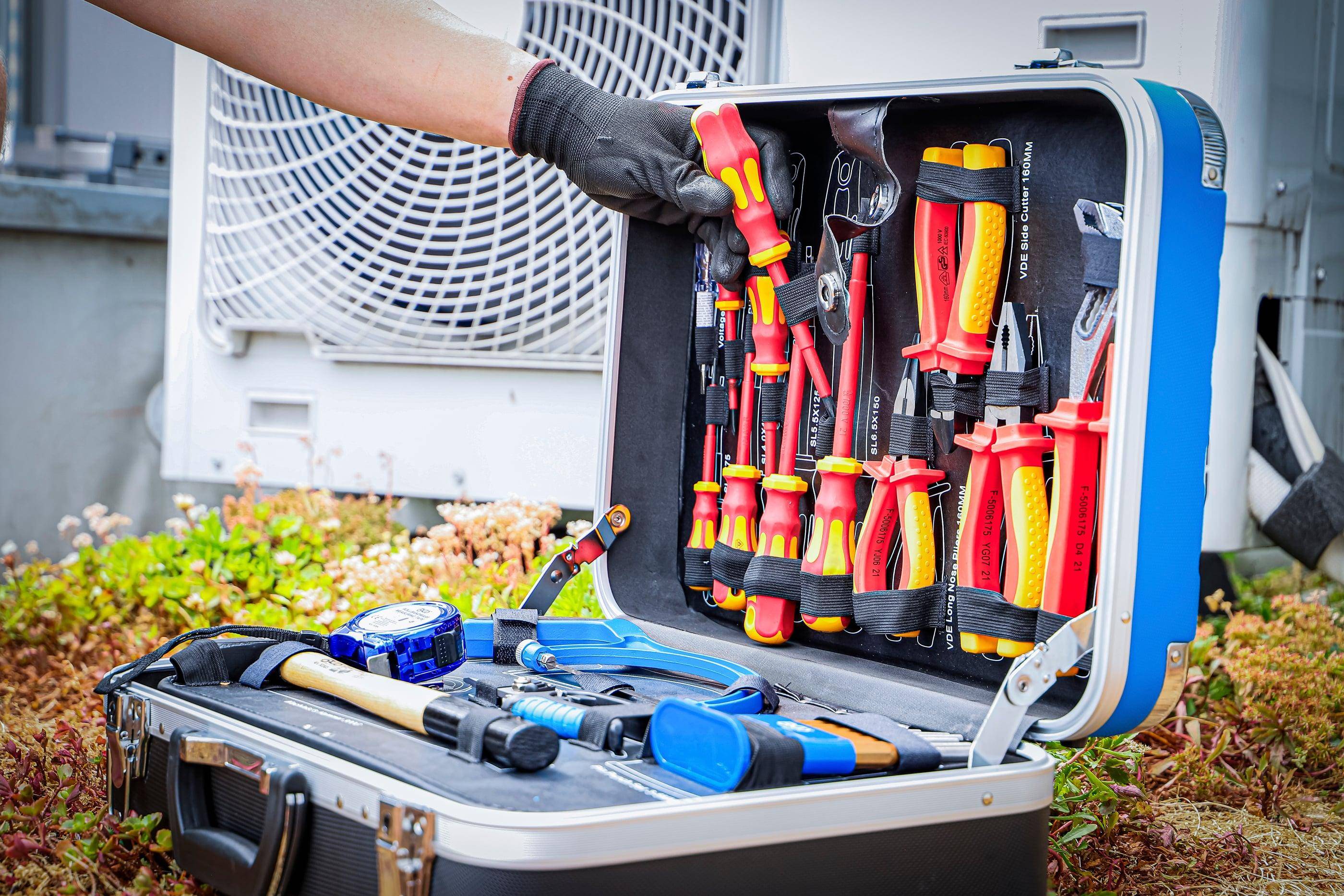 Eine Person mit Handschuhen öffnet eine Werkzeugkiste voller verschiedener isolierter Schraubenzieher und Werkzeuge in der Nähe von Klimaanlagen im Freien.