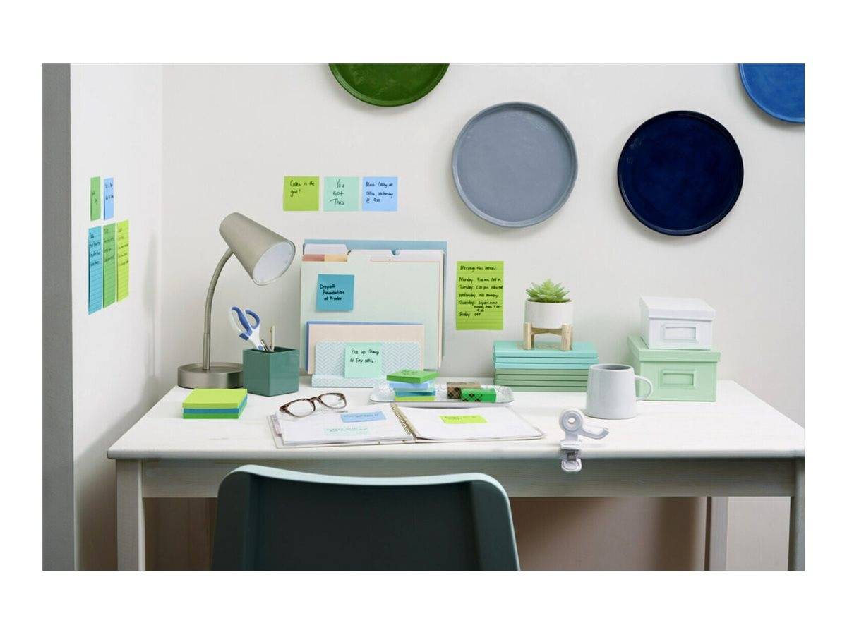 Organisierter Schreibtischarbeitsplatz mit Lampe, Notizbüchern, Haftnotizen, Brille, kleiner Pflanze und an der Wand angebrachten Tellern in Blau- und Grüntönen.