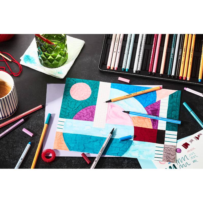 Künstlerbedarf auf einem Tisch mit einer farbenfrohen geometrischen Zeichnung, Stiften, Bleistiften, einem grünen Glasstifthalter und einer gestreiften Tasse.