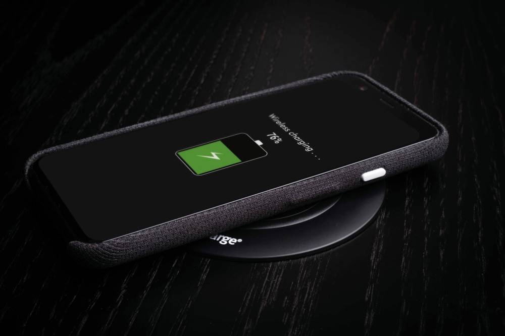 Ein Smartphone, das drahtlos auf einer Ladestation aufgeladen wird, mit einem Bildschirm, der ein Batteriesymbol und den Text ‚Drahtloses Laden: 76%' auf einer dunklen Oberfläche zeigt.