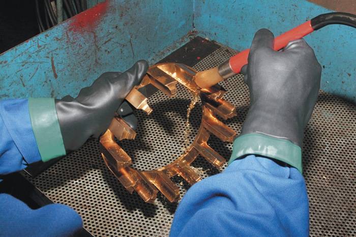 Eine Person mit Schutzhandschuhen und einem blauen Mantel benutzt ein Werkzeug, um eine Kupferkomponente in einem industriellen Umfeld zu montieren oder zu justieren.