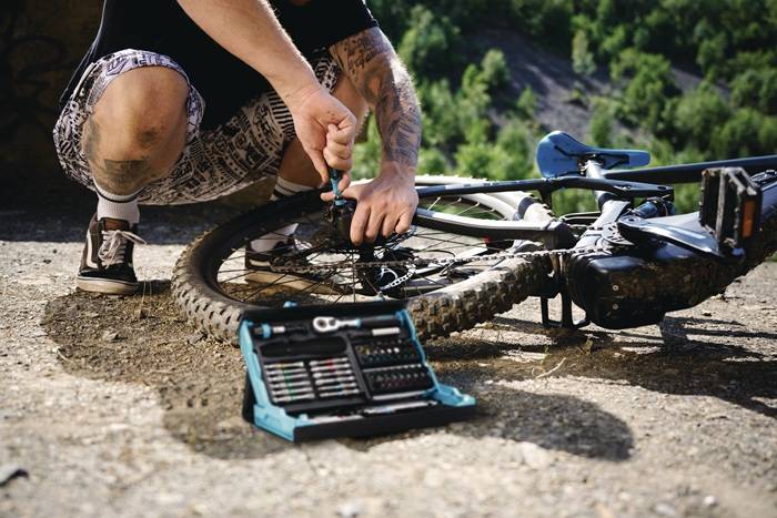 Eine Person repariert einen Fahrradreifen auf einem felsigen Pfad, umgeben von grünen Bäumen, mit einem geöffneten Werkzeugkasten in der Nähe.