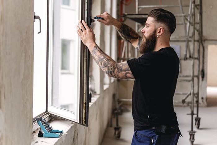 Eine Person mit Bart und Tätowierungen montiert einen Fensterrahmen in einem renovierungsbedürftigen Gebäude und konzentriert sich dabei auf Präzision und Ausrichtung.