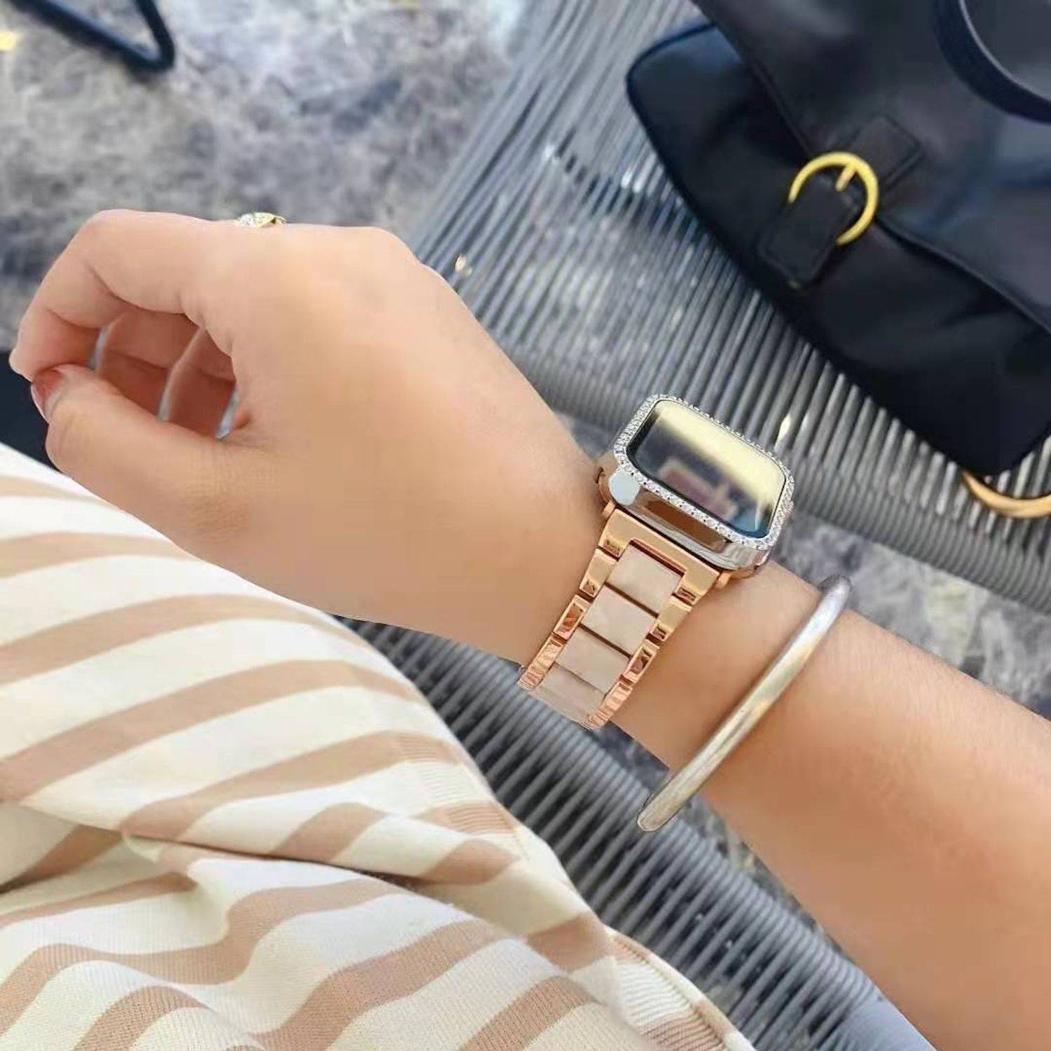 Nahaufnahme des Handgelenks einer Person, die eine Smartwatch in Roségold und ein silbernes Armband trägt, mit einer schwarzen Handtasche im Hintergrund.