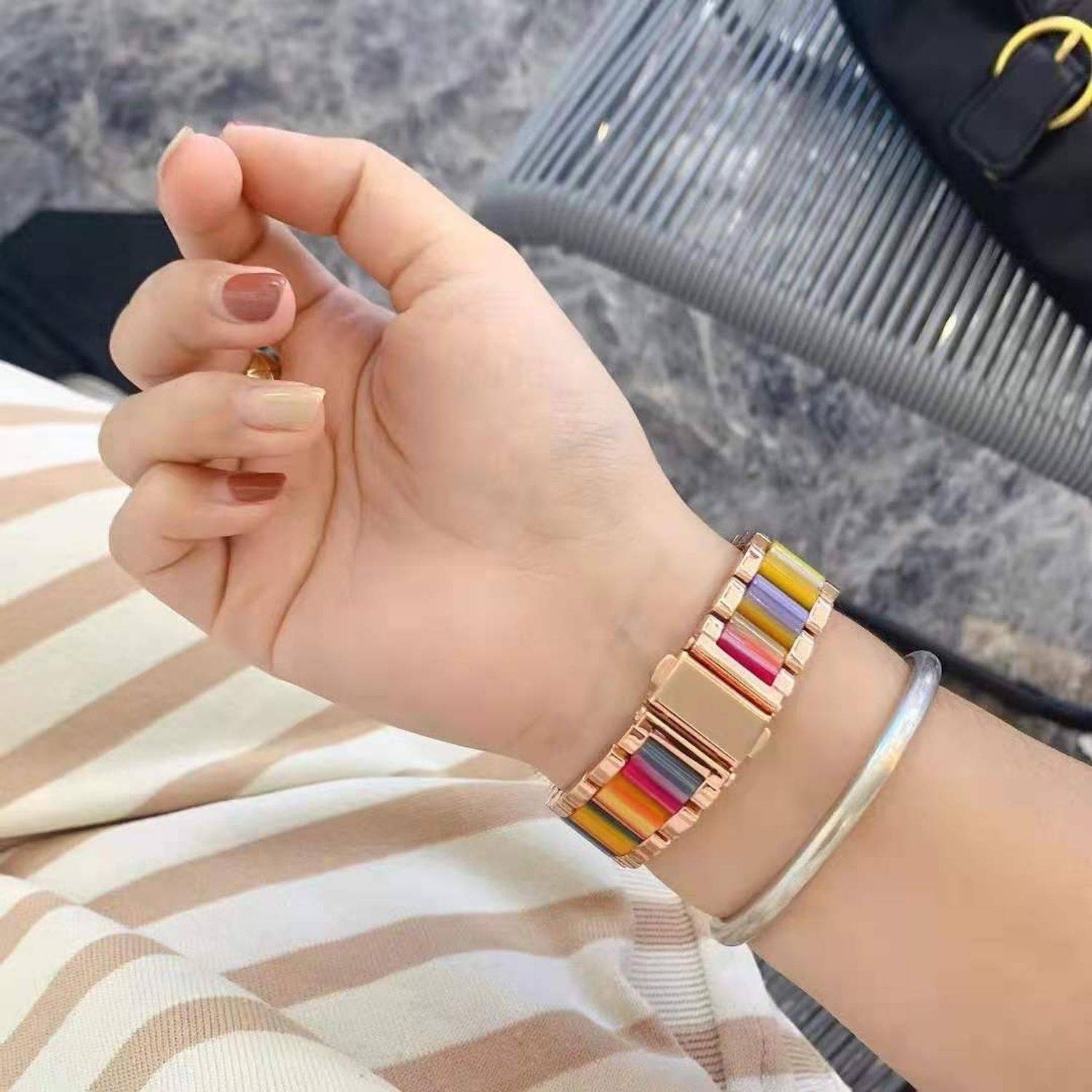 Nahaufnahme einer Frauenhand mit einer regenbogenfarbigen gestreiften Metallarmbanduhr und einem silbernen Armreif.