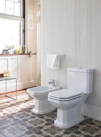 Ein heller Badezimmer mit einem Fenster, ausgestattet mit einer Toilette, einem Bidet und einer Handtuchstange auf einem gefliesten Boden. Ein Regal trägt aufgerollte Handtücher und Flaschen.
