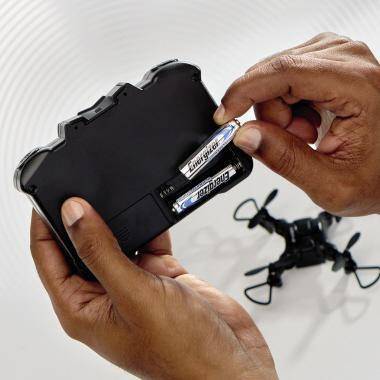 Hände führen Batterien in eine kleine schwarze Drohnen-Fernbedienung ein, während die Drohne in der Nähe auf einer weißen Oberfläche sitzt.