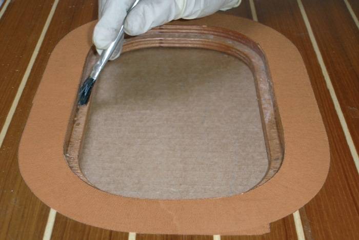 Eine Hand mit Handschuh verwendet einen Pinsel, um Kleber in eine flache, rechteckige Holzvertiefung auf einem Tisch aufzutragen, höchstwahrscheinlich für eine Montagetätigkeit.