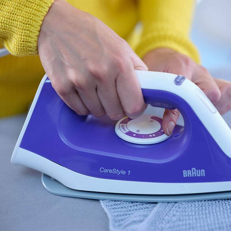 Hand, die ein Braun CareStyle 1 Dampfbügeleisen auf einem blauen Kleidungsstück verwendet, wobei Stoff auf einer ebenen Fläche gebügelt wird.