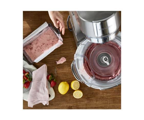 Overhead-Ansicht eines Mixers, der eine rosa Erdbeer-Mischung auf einer Holztheke zubereitet, mit einer Hand, die Zutaten hinzufügt, Erdbeeren, Zitronen und ein Handtuch in der Nähe.