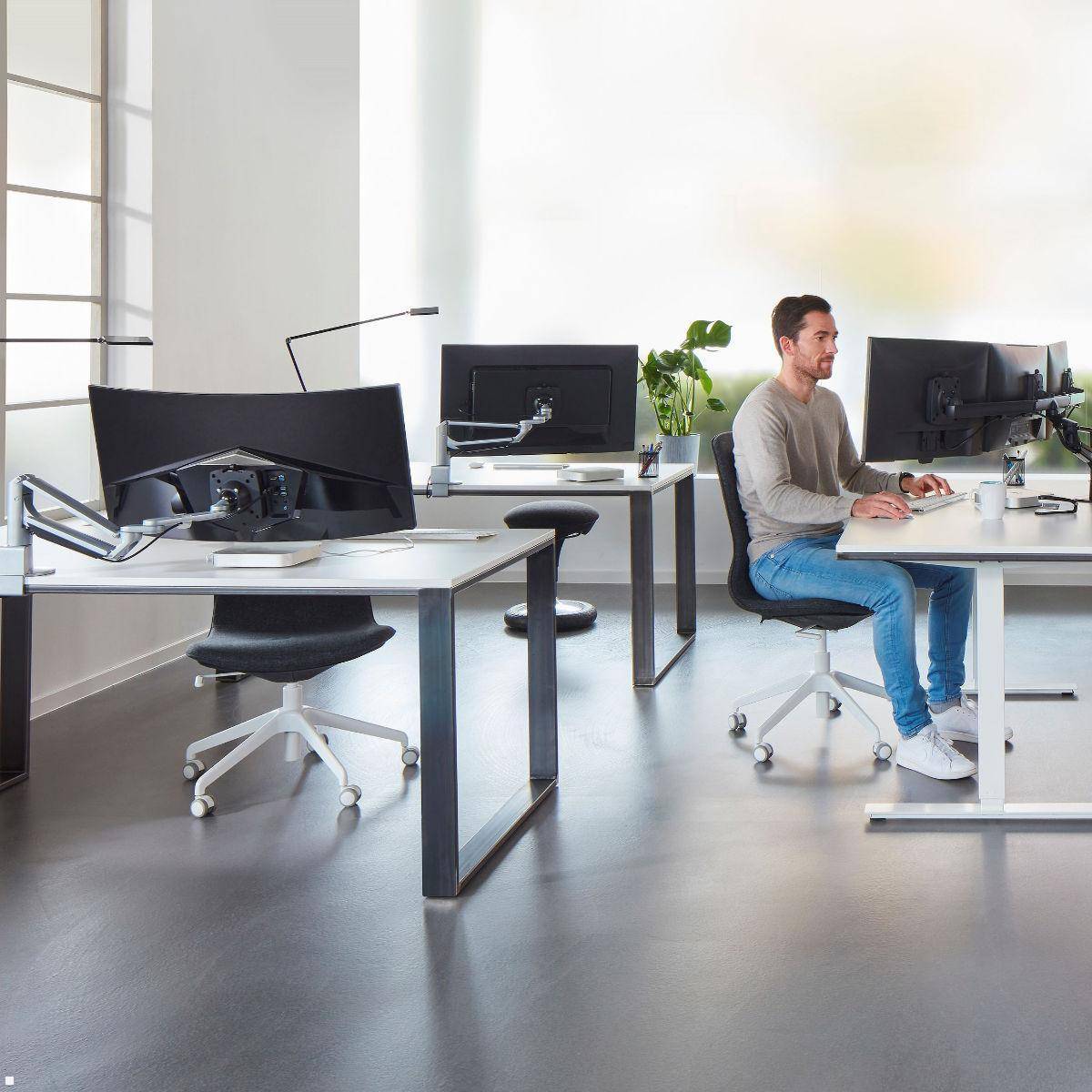 Eine Person, die an einem modernen Schreibtisch in einem offenen Büroraum sitzt und an einem Computer arbeitet. Der Raum ist hell und enthält mehrere Schreibtische und Monitore.