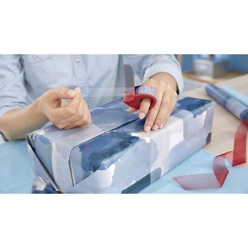 Eine Person, die ein Geschenk mit blau gemustertem Papier einwickelt und dabei einen Klebebandabroller verwendet und eine Klebestreifenrolle in der Hand hält, an einem Tisch mit rotem Band in der Nähe.