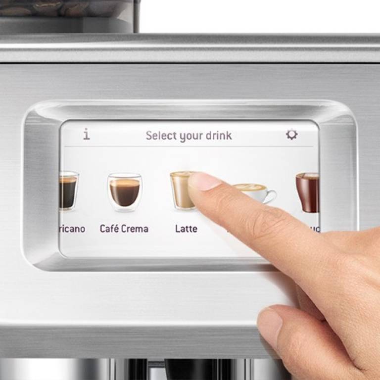 Eine Hand tippt auf den Touchscreen eines Kaffeeautomaten, um ein Getränk auszuwählen, wobei Optionen wie "Café Crema"" und ""Latte"" sichtbar sind.