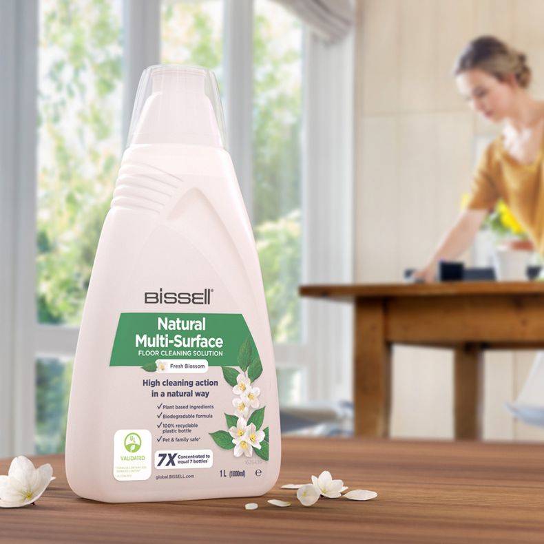 Bissell Natural Multi-Surface Bodenreinigungslösung Flasche auf einem Holztisch, mit einer Frau im Hintergrund beim Reinigen und weißen Blumen rundherum.