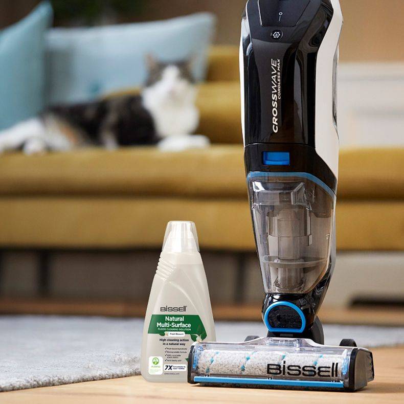 Bissell CrossWave Bodeneinigers mit Reinigungslösungsflasche auf einem Teppich in einem Wohnzimmer, mit einer unscharfen Katze auf dem Sofa im Hintergrund.