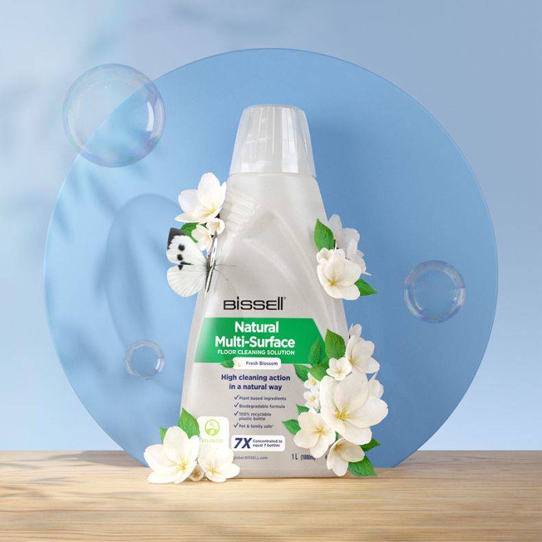 Bissell Natural Multi-Surface Bodenreinigungslösungsflasche mit weißen Blumen und Schmetterlingen, dargestellt auf einer Holzoberfläche vor einem blauen kreisförmigen Hintergrund.