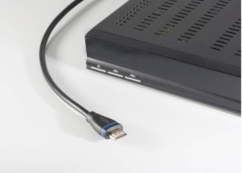 Nahaufnahme eines HDMI-Kabelsteckers neben einer schwarzen Set-Top-Box oder eines Mediaplayers auf einer weißen Oberfläche.