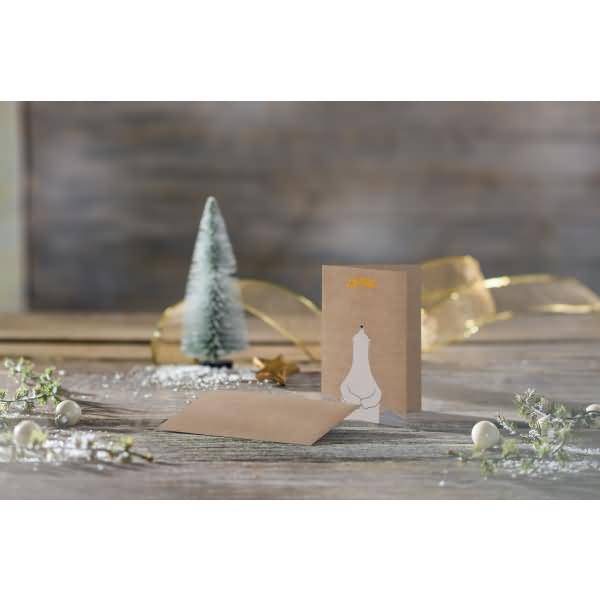Weihnachtsgrußkarte auf einem rustikalen Tisch mit einem kleinen gefrosteten Baum, goldenem Band und Grünwerk.