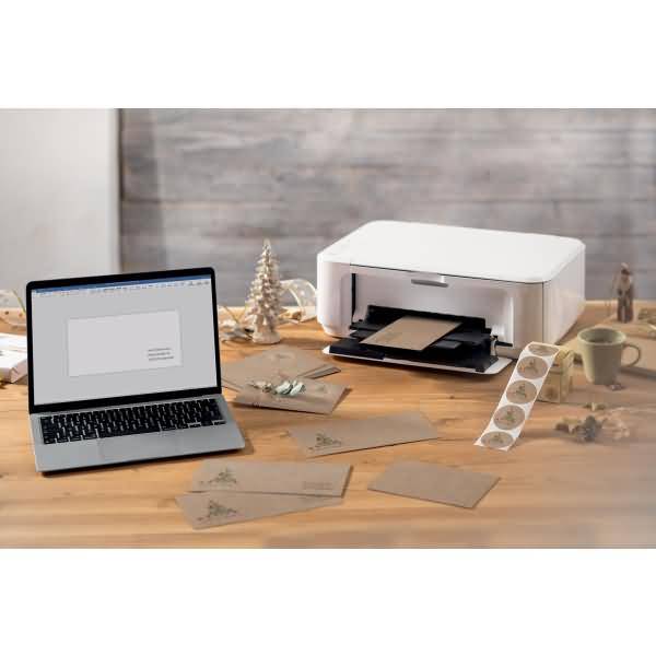 Laptop und Drucker auf einem Basteltisch mit Umschlägen, Papier und Klebeband-Aufklebern, was auf den Heimdruck von personalisierten Karten oder Einladungen hindeutet.