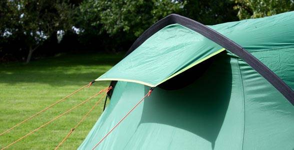 Nahaufnahme eines grünen Campingzelts, das auf Gras im Freien aufgespannt ist, mit gespannten Abspannseilen und Bäumen im Hintergrund.